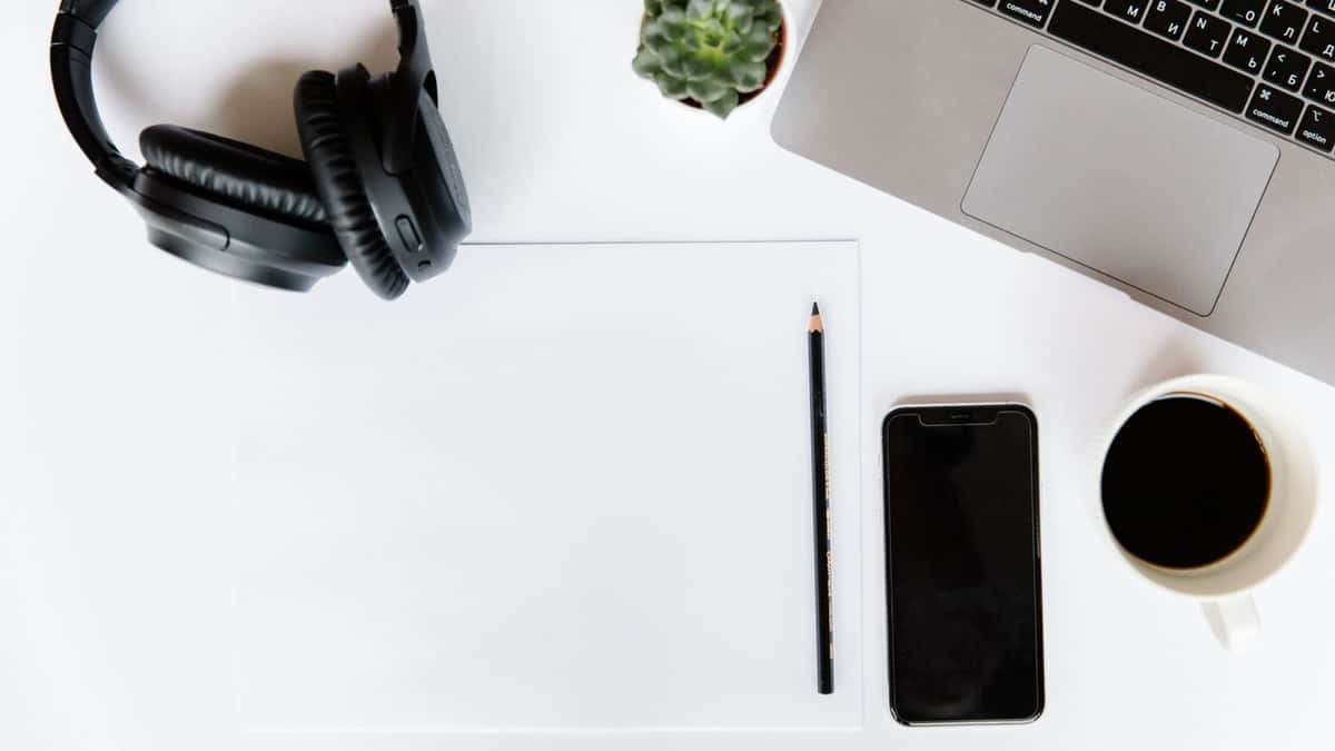 Escritorio blanco con audífonos, teléfono, libreta, lápiz, portátil y taza de café